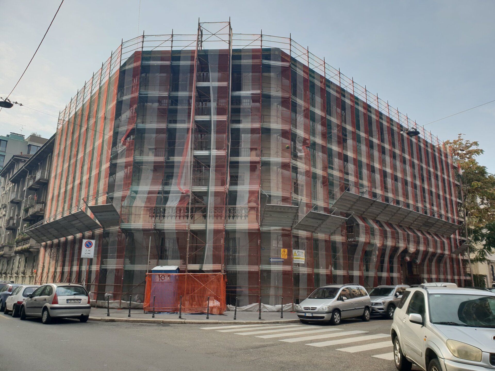 Edificio in fase di ristrutturazione, coperto da impalcature e reti di sicurezza, all'angolo di una strada con auto parcheggiate.