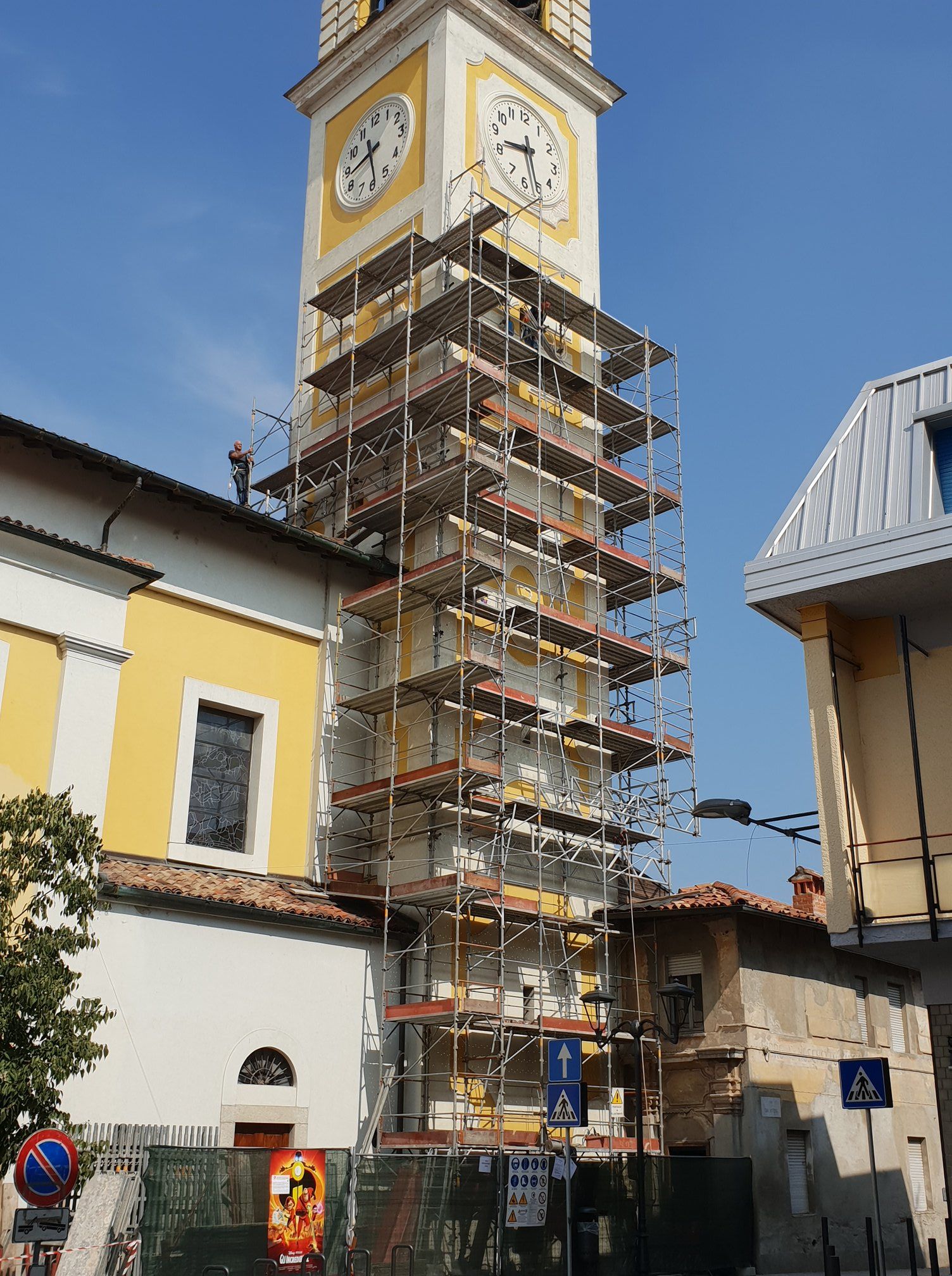 Campanile della chiesa con impalcature per il restauro; edifici gialli, bianchi e marroni contro un cielo blu.