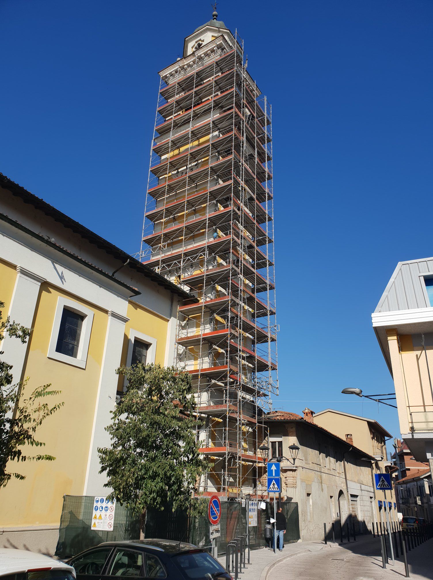 Alto campanile di una chiesa sotto le impalcature per lavori di ristrutturazione, in una strada con edifici, cielo azzurro e terso.