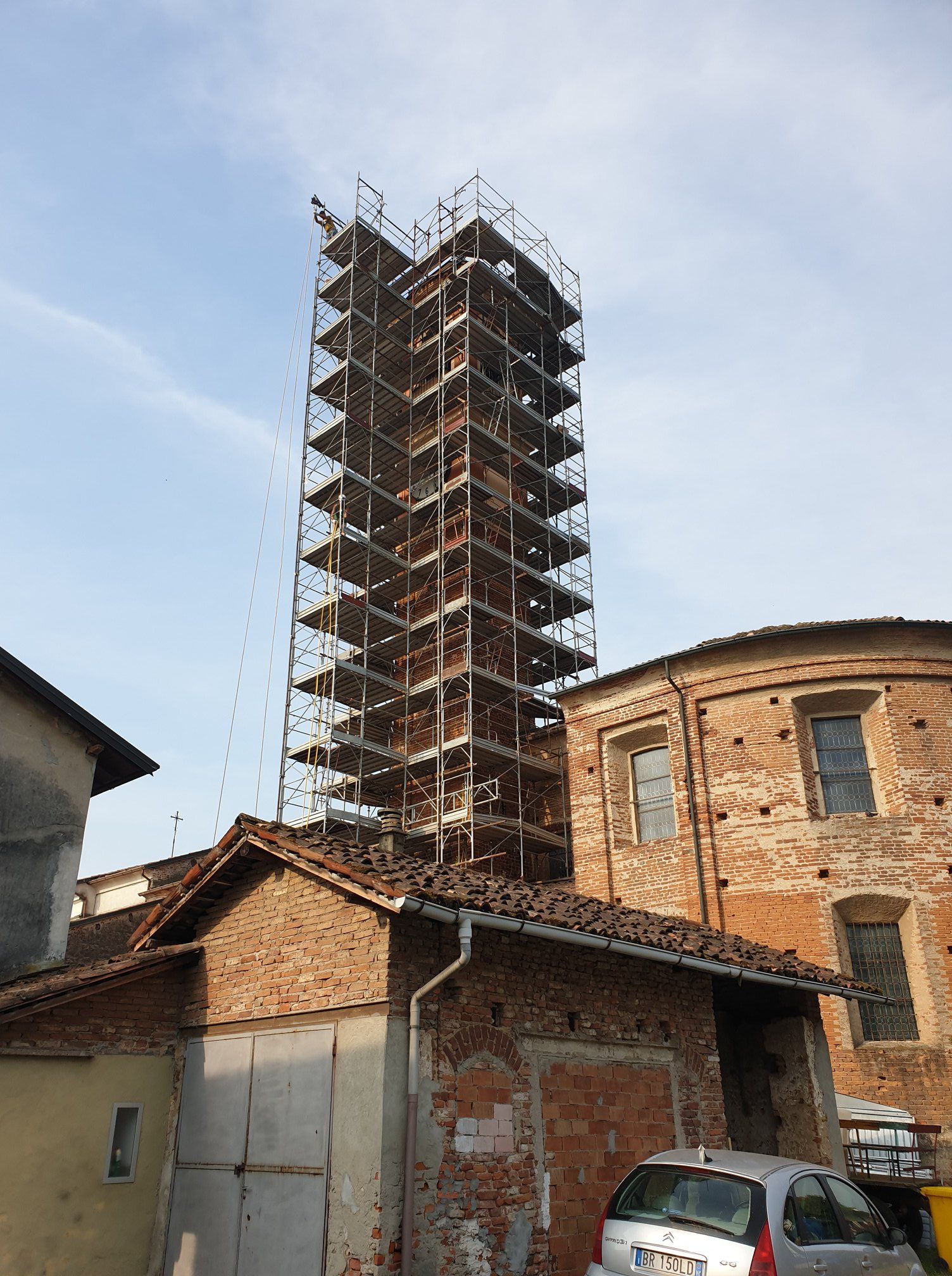 Alta torre in costruzione con impalcature, contro un cielo azzurro, circondata da edifici.