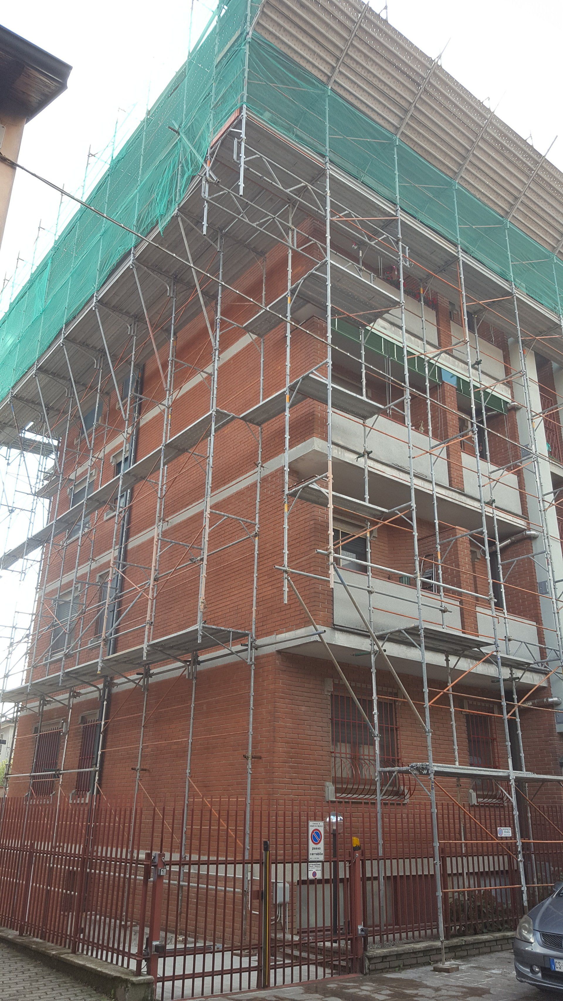 Facciata dell'edificio in costruzione, esterno in mattoni rossi, impalcature e rete verde.