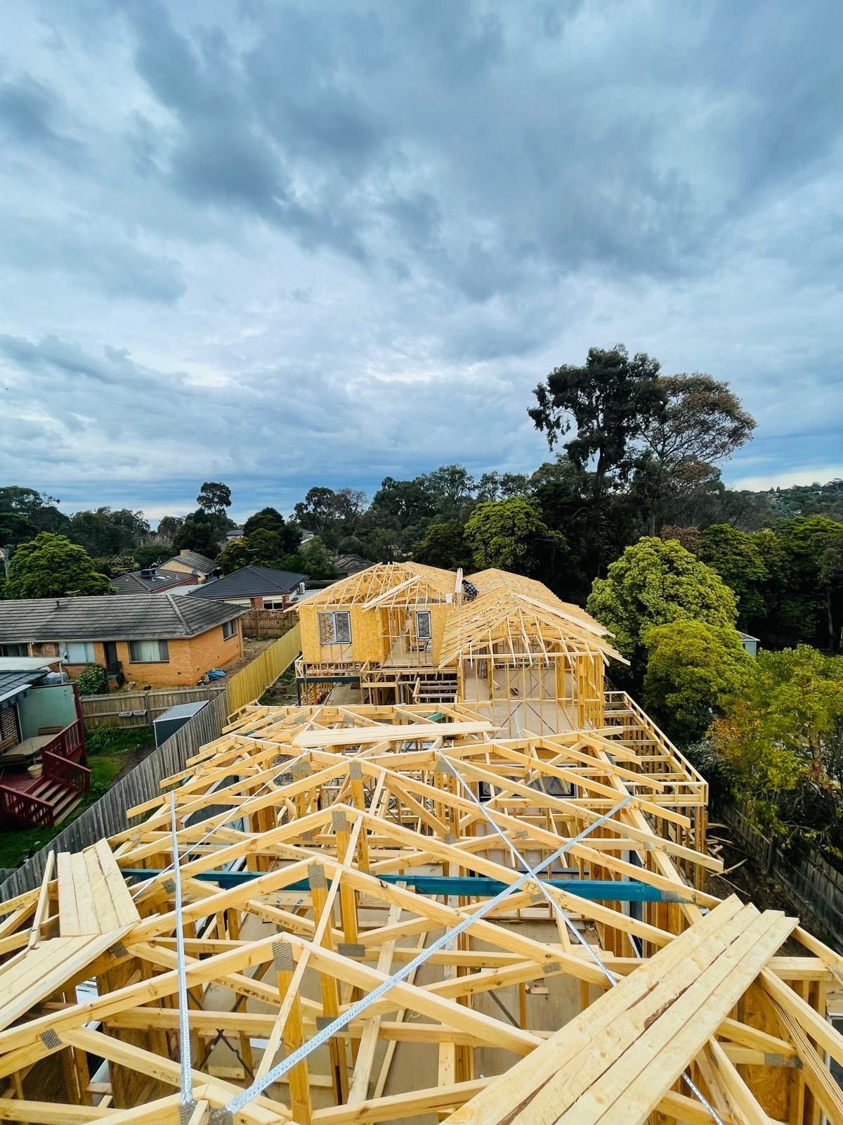 Wooden framework of a house under construction, viewed from above, with cloudy sky and suburban background.