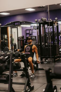 Man seated on a gym machine in a purple-lit fitness center, with dumbbells and cable equipment nearby.