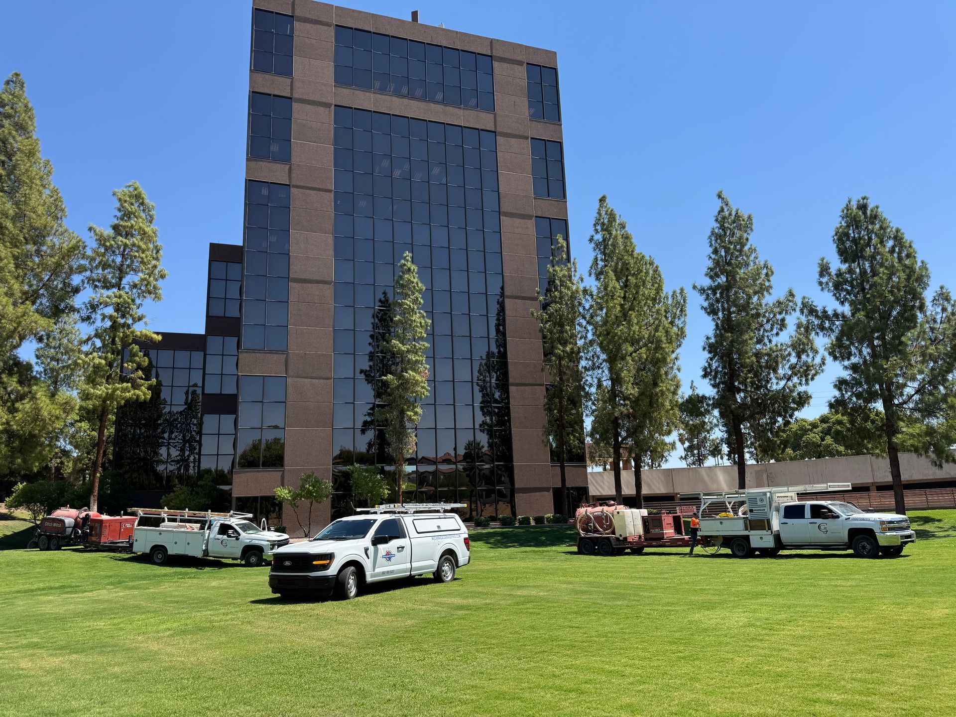 Several work trucks are parked on a green lawn in front of a tall, modern office building with dark, reflective windows.