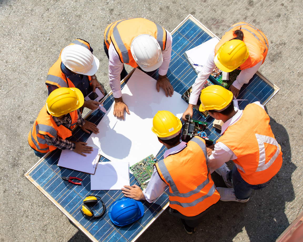 Top-down view of six professionals in orange safety vests and hard hats reviewing architectural plans on a solar panel.