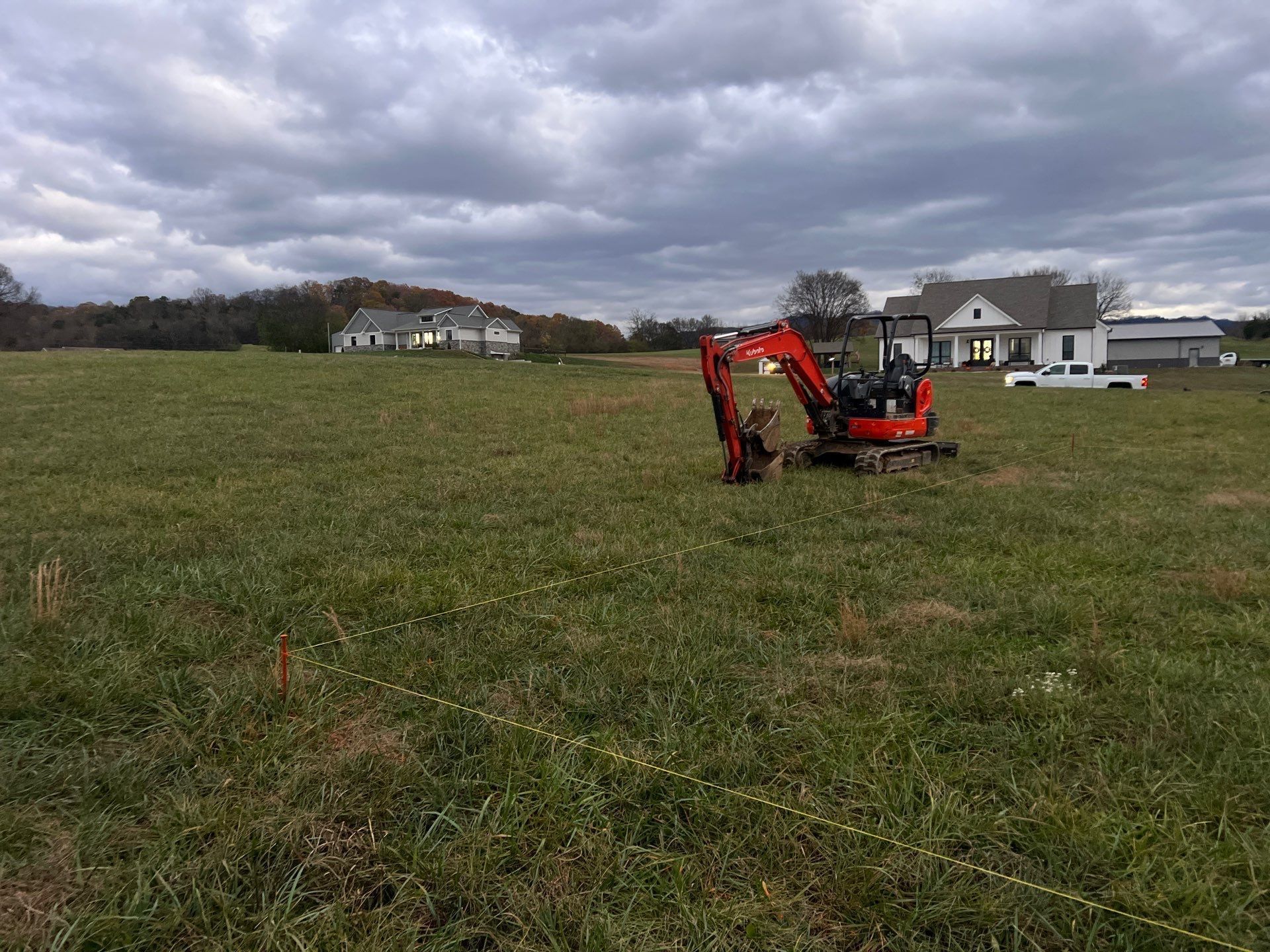 A small red excavator is sitting in a grassy field in front of a house.