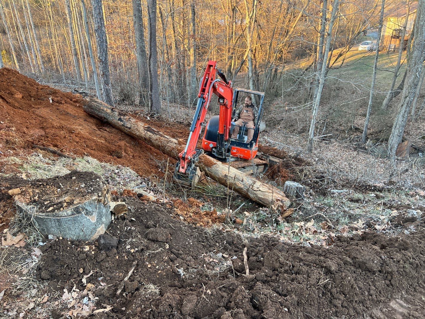 A red excavator is digging a hole in the dirt in the woods.