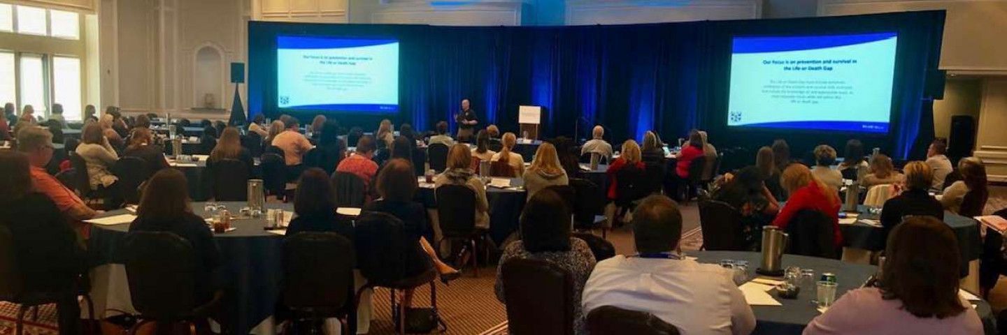 Audience at a conference in a large room, watching a presentation on a screen.