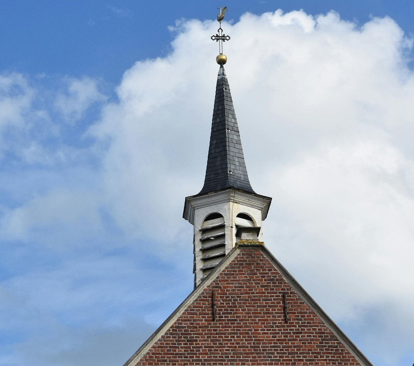 Brick church roof with steeple, topped by a cross and weather vane, against a cloudy blue sky.