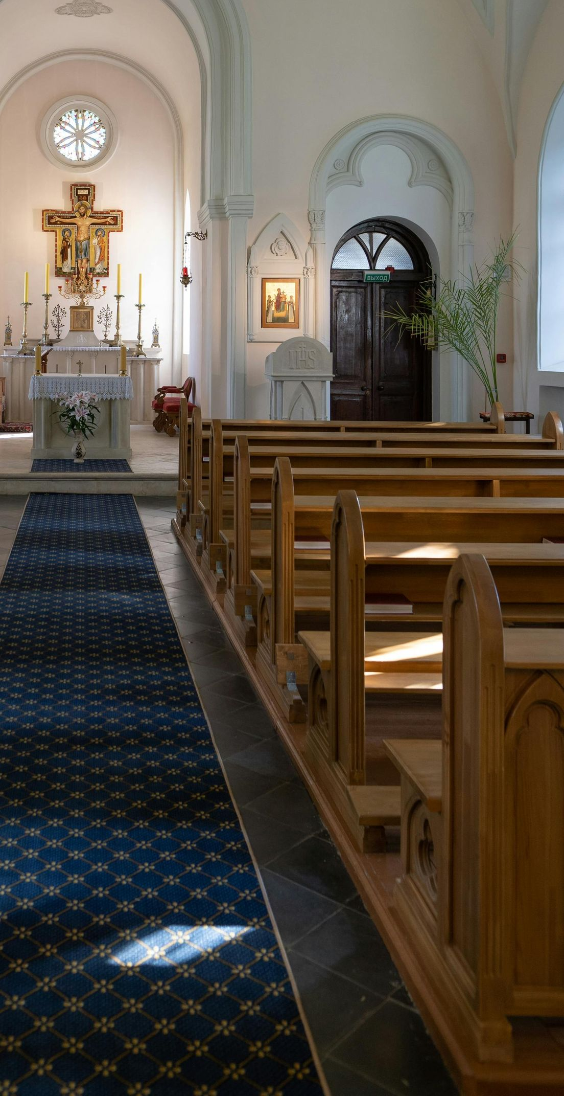 Interior view of a church. Wooden pews line the right side, a blue carpeted aisle leads to the altar and crucifix.