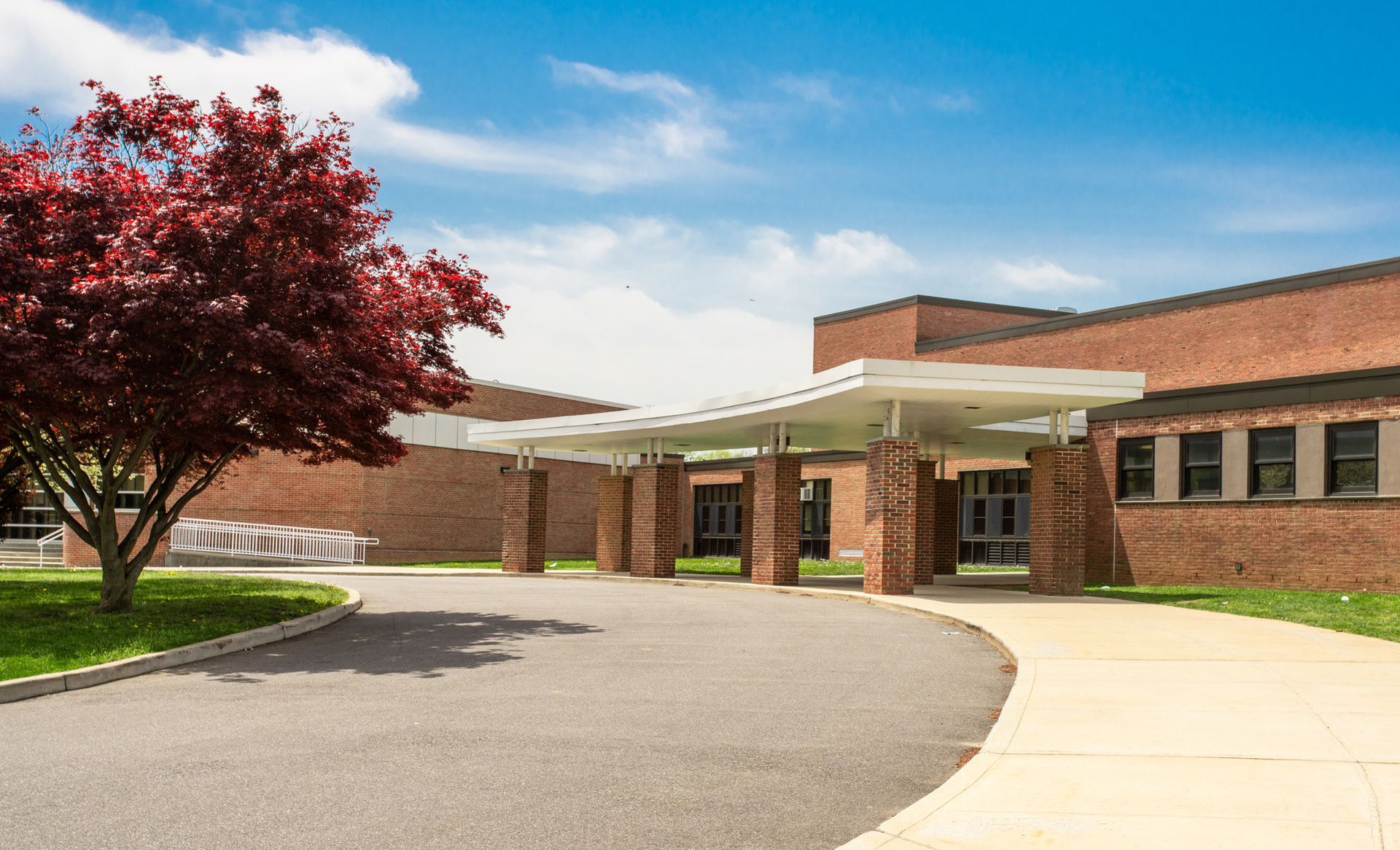Red brick building with covered entrance, paved driveway, and red tree.