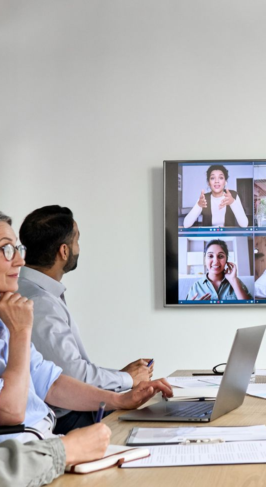 People in meeting at table with laptop, video conference on screen.