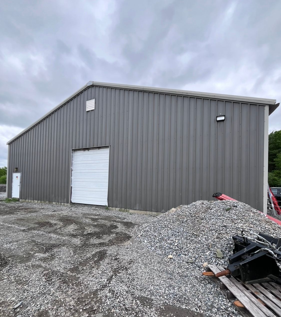 A large gray metal building with a white garage door is sitting next to a pile of gravel.