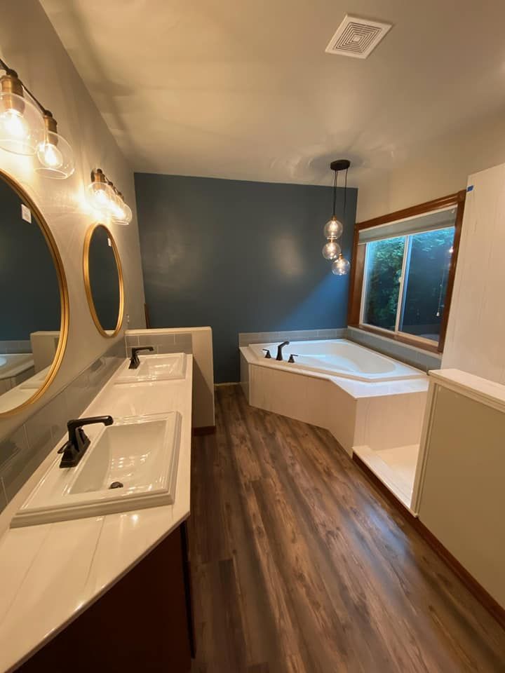 Bathroom with dual sinks, a tub, and wood-look flooring. Blue accent wall, gold-framed mirrors, and black fixtures.