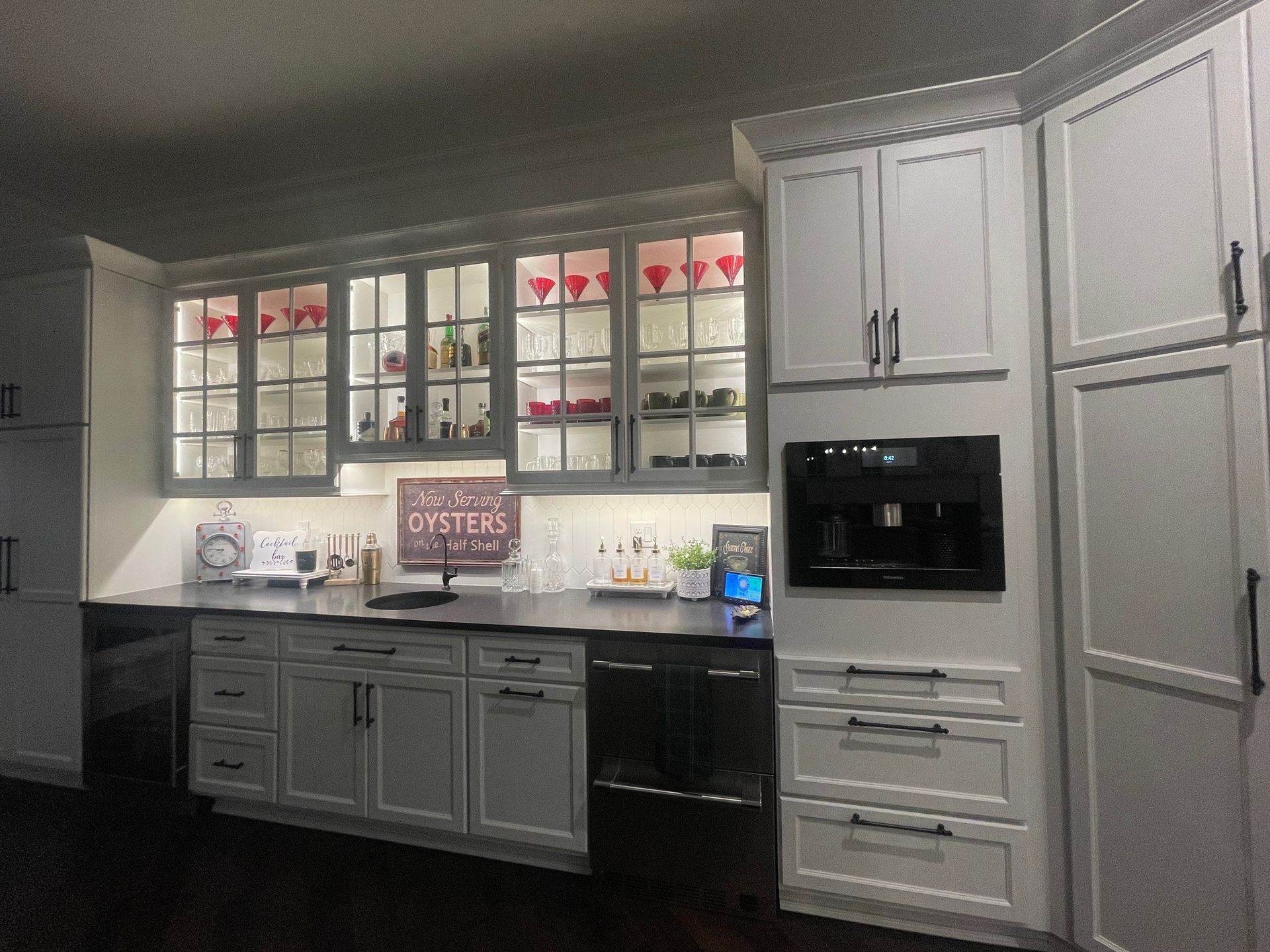 Kitchen with white cabinets, glass-front upper cabinets with lit interiors, a built-in coffee machine, and dark countertops.
