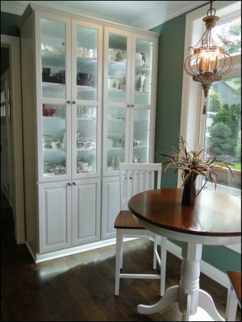White built-in cabinet with glass doors displaying dishware.  Table and chair in front, next to a window.