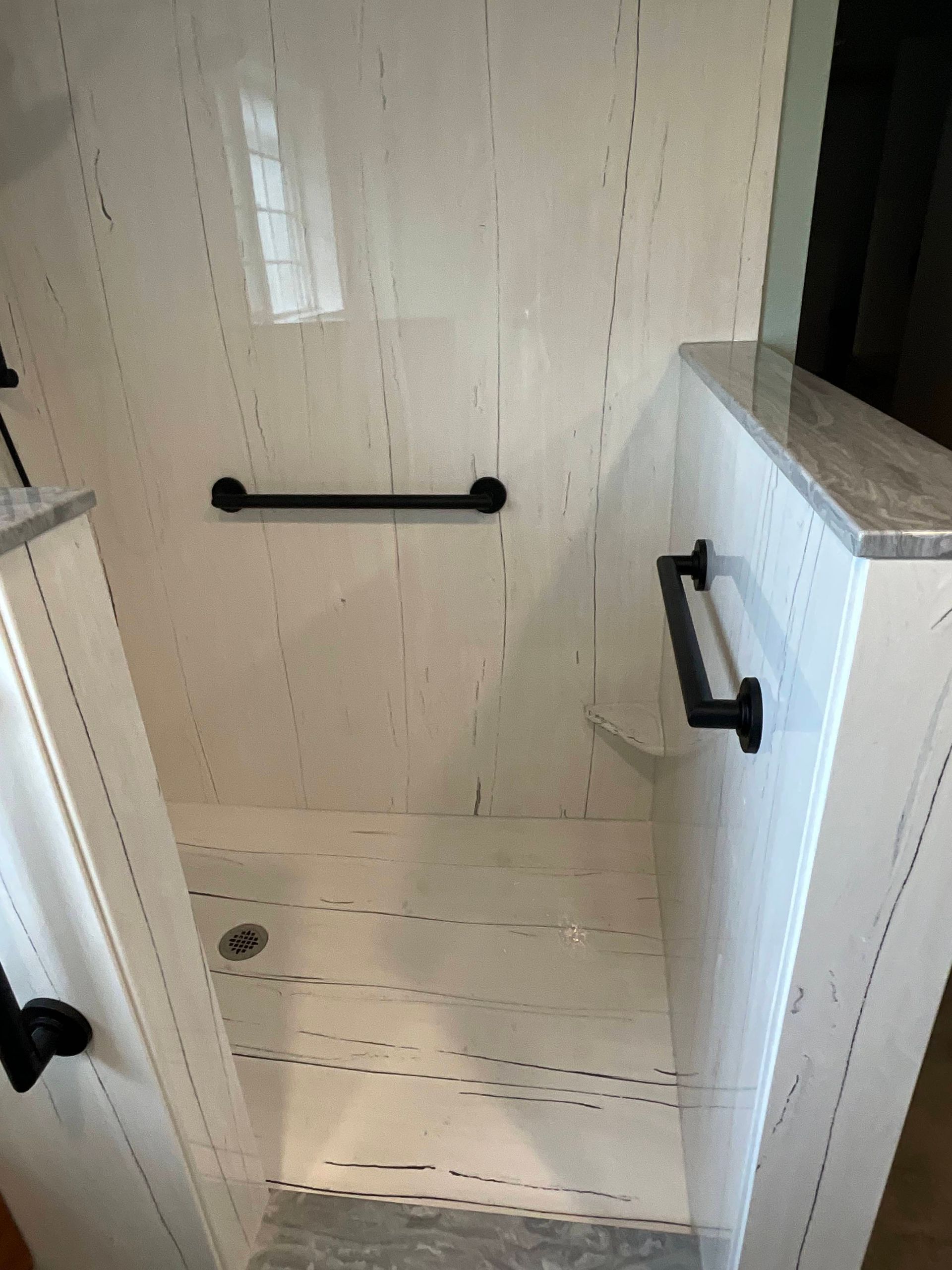 Shower stall with white paneled walls, black handrails, and a glass door.