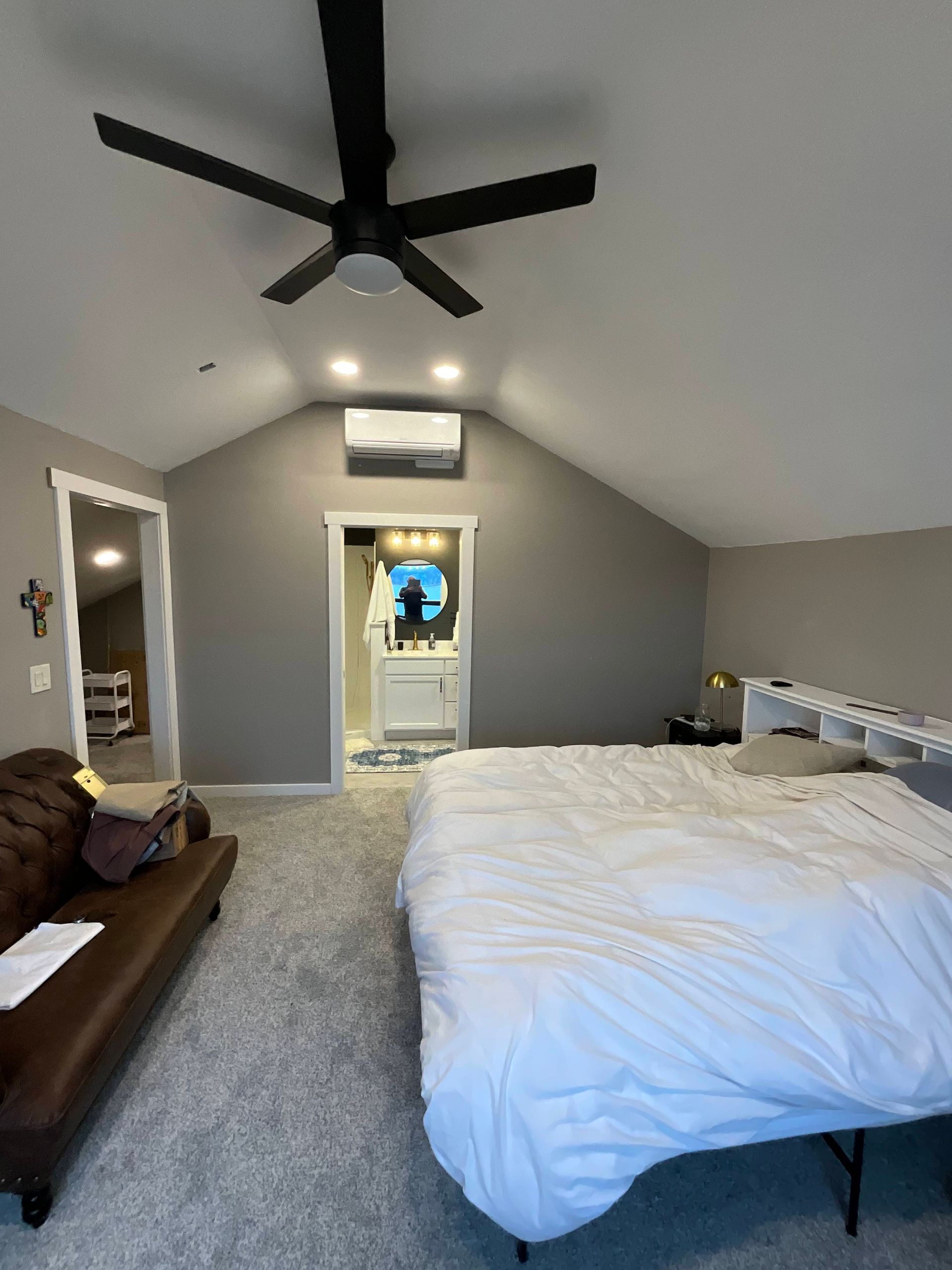 Bedroom with bed, couch, and a view to the bathroom. Gray walls, ceiling fan, and carpet.