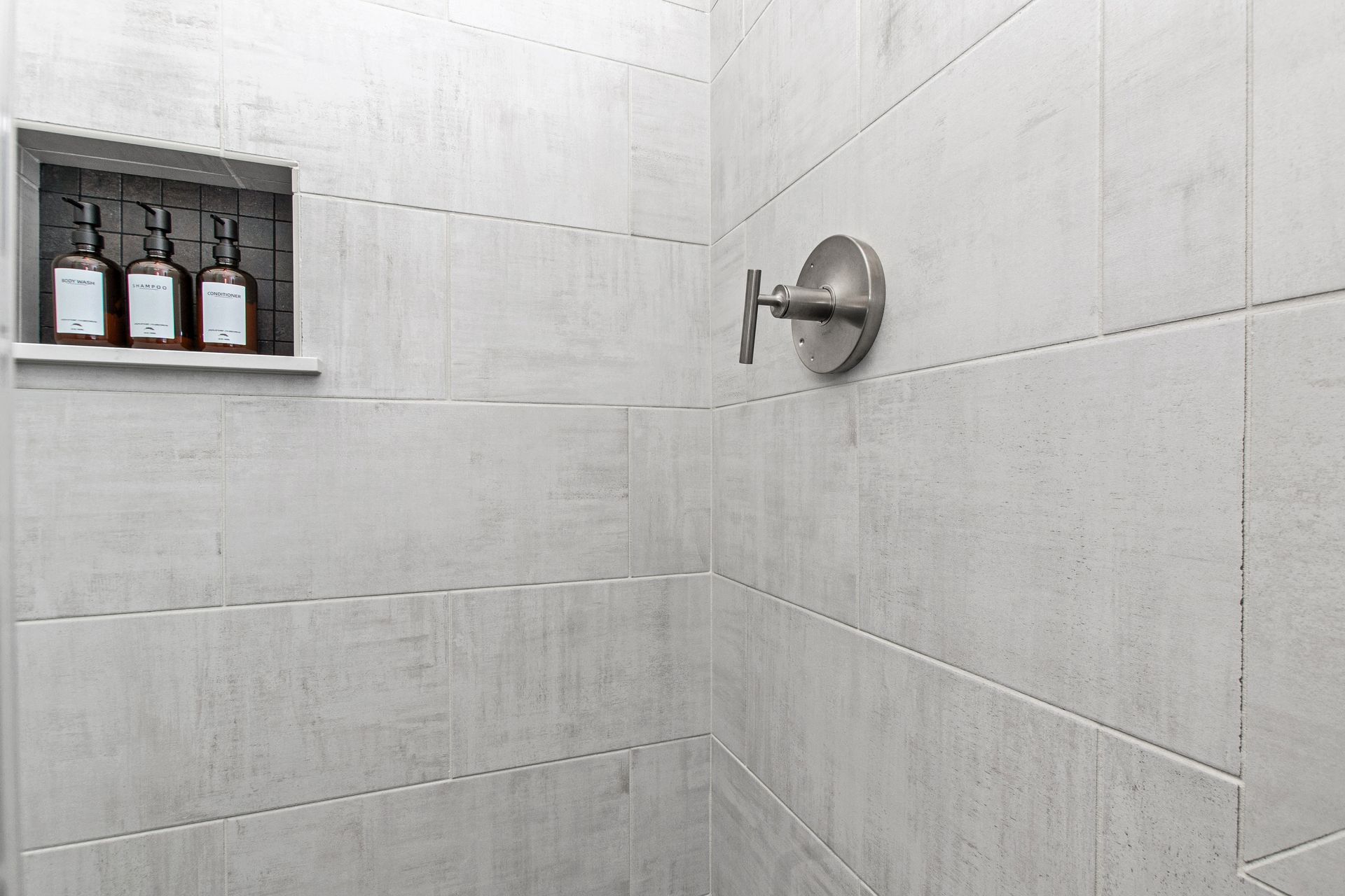 A shower with white tiles and a stainless steel shower head.