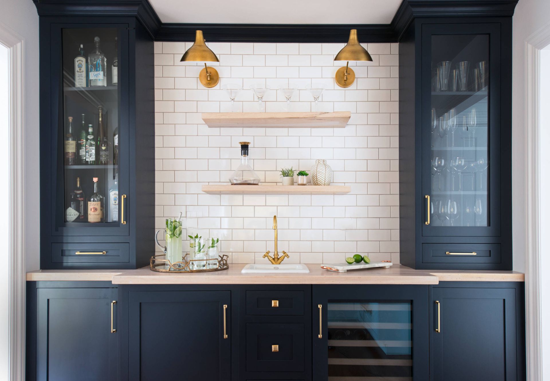 Navy blue home bar with brass fixtures and white brick backsplash.