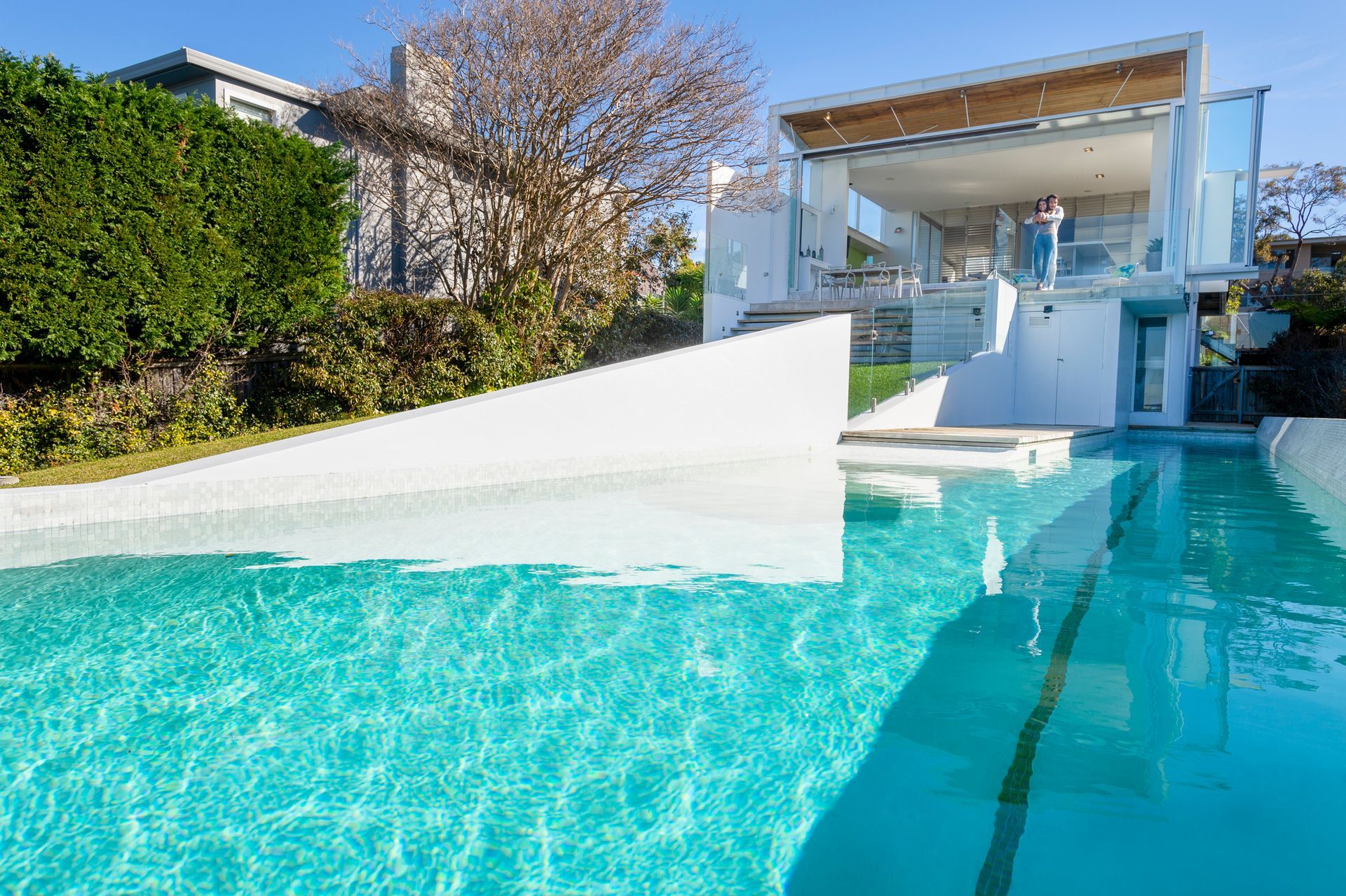 Modern luxury home with glass windows, balcony, and pool surrounded by greenery in daylight.