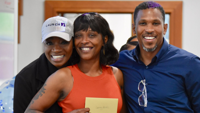 Three smiling people indoors, one holding a yellow envelope, standing close together.