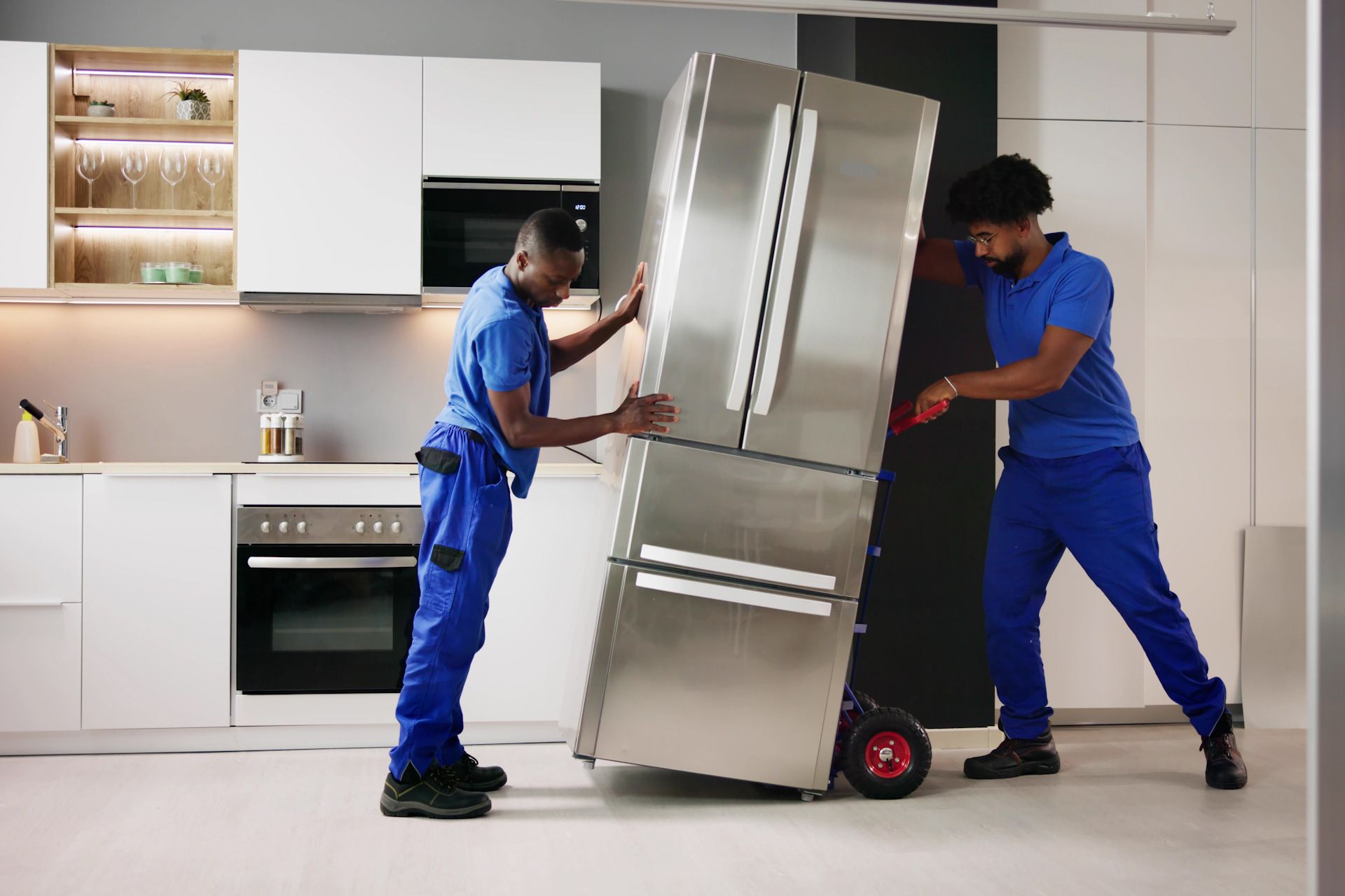 Two men are carrying a refrigerator on a dolly in a kitchen.