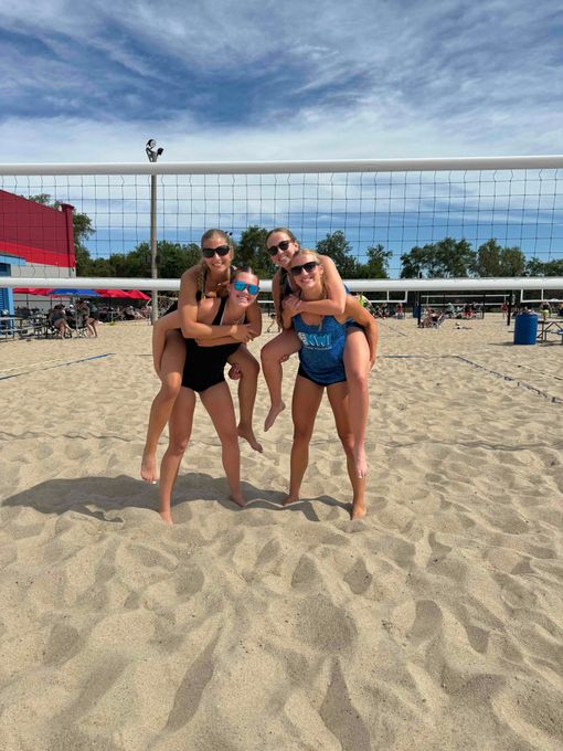 Three women are posing for a picture on a beach with a volleyball net in the background.