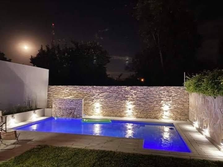 Vista nocturna de una piscina iluminada con muro de piedra y paisajismo. El agua azul refleja la luz.