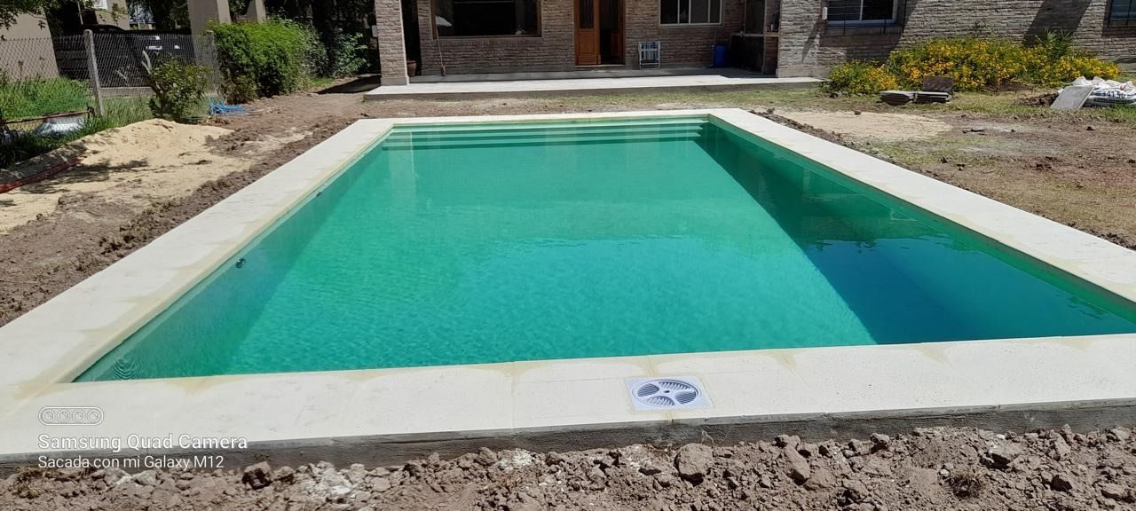 Piscina rectangular de agua turquesa, rodeada de hormigón y tierra. Al fondo se ve una casa.