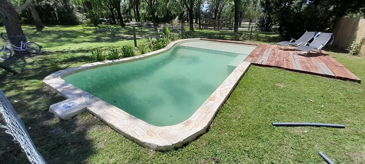 Una piscina rodeada de césped verde, con tumbonas en una terraza de madera. Árboles al fondo.