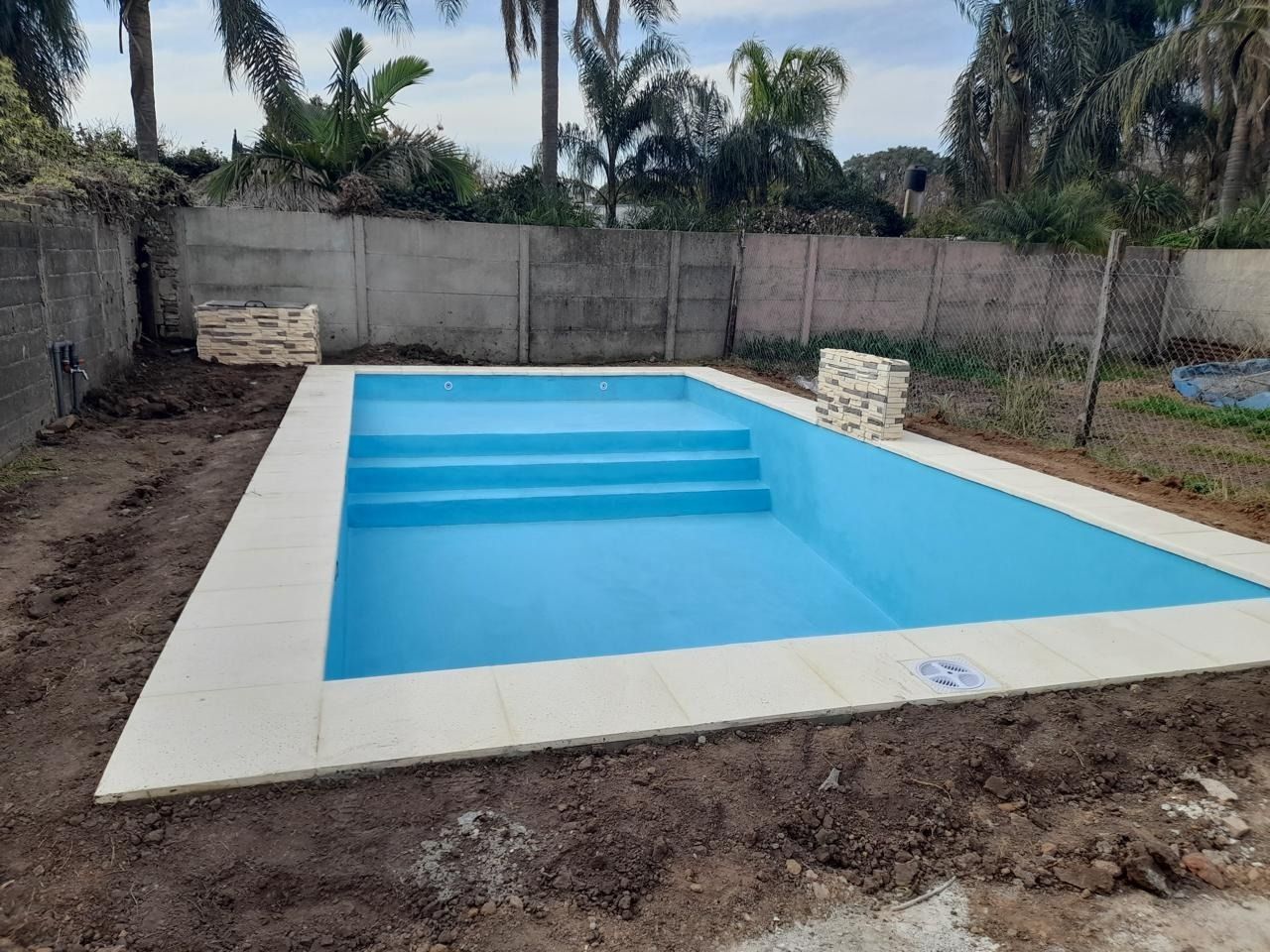 Una piscina rectangular de agua azul y borde claro. Rodeada de tierra y un muro de hormigón.