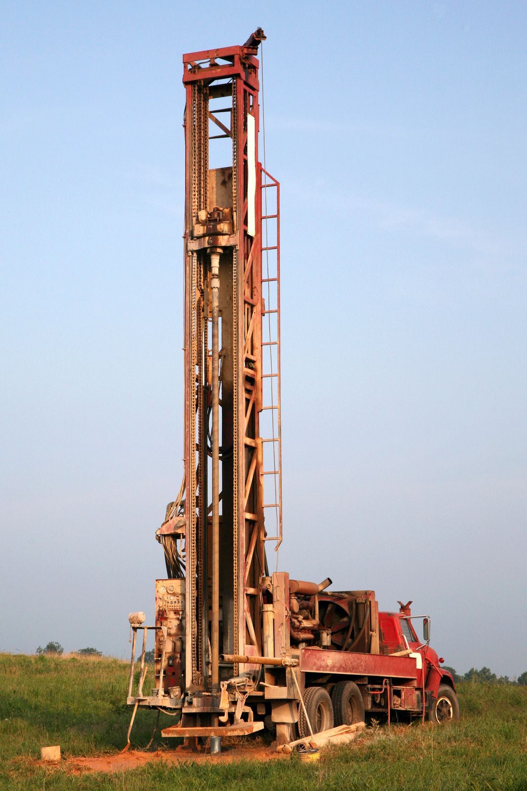 A red truck-mounted drill rig stands upright in a grassy field against a light blue sky.