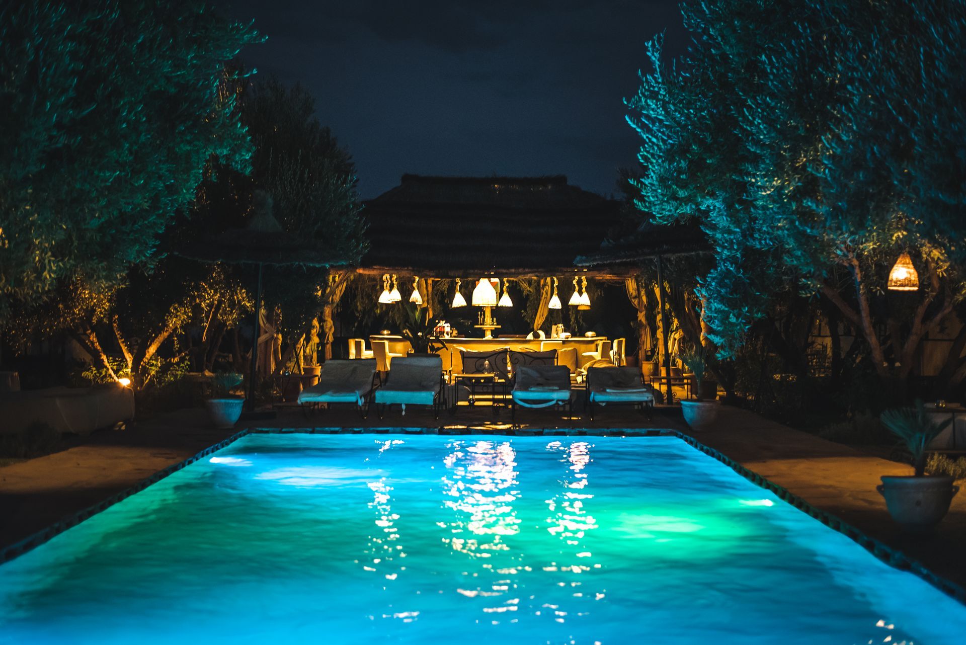Night-lit backyard pool with turquoise water, surrounded by trees and a warmly lit pavilion.