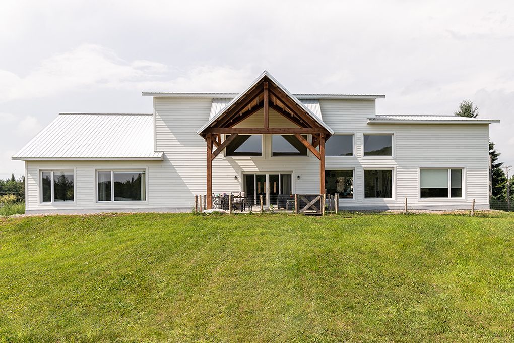 Une grande maison blanche avec un porche en bois se trouve au sommet d'un champ verdoyant.