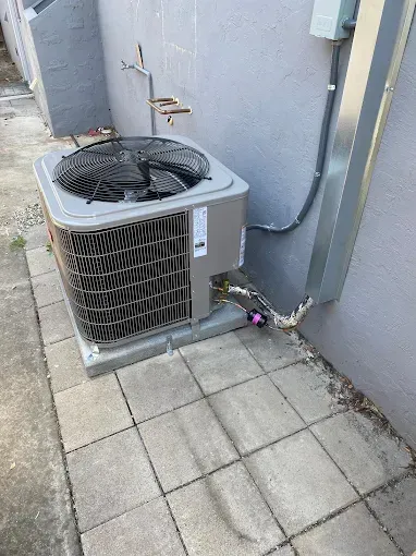 An outdoor gray HVAC condenser unit sits on a concrete patio next to a gray wall with an attached metal conduit.