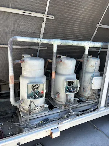 Three industrial HVAC compressors in a row on a rooftop unit with exposed electrical terminal boxes and piping.