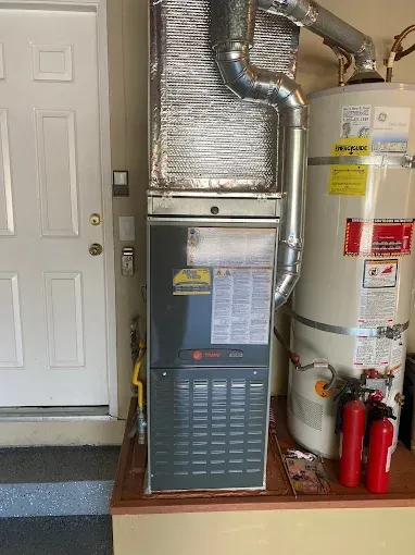A Trane gas furnace and a water heater installed side-by-side on an elevated platform in a garage near a door.