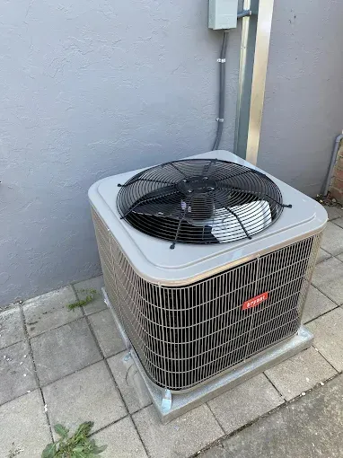 A gray residential air conditioning condenser unit sits on a concrete patio against a gray wall.