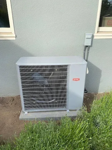 A gray Bryant air conditioning unit sits on a concrete pad against an exterior wall near green landscaping.