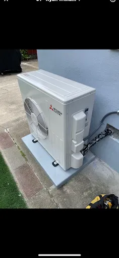 A white Mitsubishi outdoor heat pump unit mounted on a grey concrete base next to a wall on a paved patio.