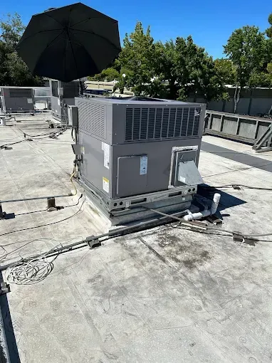A rooftop HVAC unit on a flat gray roof under a clear blue sky, with a black umbrella placed atop it.
