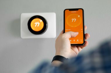 A person uses a smartphone app to adjust a wall-mounted Nest smart thermostat, both displaying an orange screen at 77°.