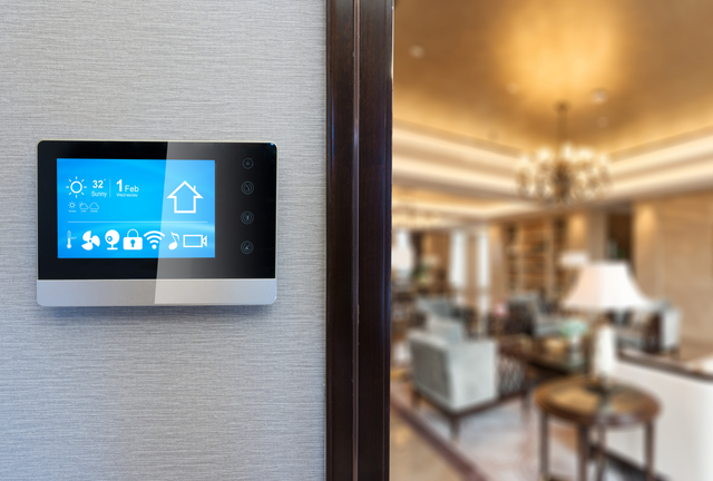 A smart home control panel mounted on a wall next to a doorway, overlooking a blurred, luxury living room interior.