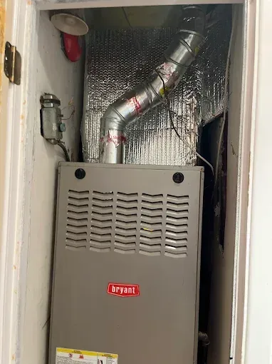 A gray Bryant HVAC furnace unit installed in a closet with a reflective insulated exhaust vent pipe above.