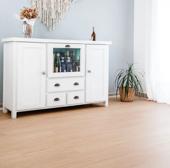 Vitrina blanca con centro de cristal. Bebidas en el interior. Flores secas, tapiz y planta. Suelo de madera clara.