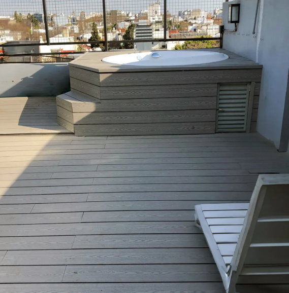 Terraza en la azotea con bañera de hidromasaje, tarima de madera y un sillón blanco, con un paisaje urbano de fondo.