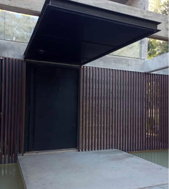 Puerta negra bajo un toldo negro. Listones verticales de madera marrón forman las paredes. Suelo de hormigón.