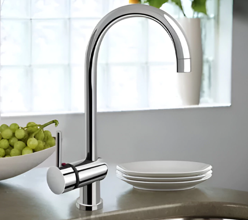 Grifo de cocina cromado de arco alto, junto a un cuenco de uvas y platos apilados.