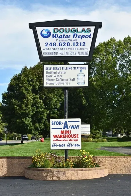 Sign for Douglas Water Depot and Stor-A-Way Mini Warehouse. White and blue signs with business info, mounted on pole, surrounded by plants.